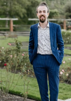 Oscar Lloyd stands in the Wellington Rose Gardens wearing a blue pinstripe suit and a floral shirt. He is smiling and has his hair styled in a bun.