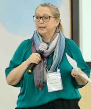 Barbara Allen holds a microphone in one hand and paper in the other while giving a talk.