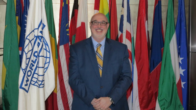 Chris is a gentleman who is standing in front of a row of flags from different nations. Man is smiling, wearing glasses and is in a dark navy blue suit.