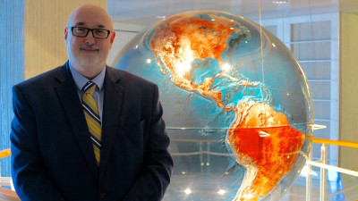 Procurement Senior Consultant Chris Browne for the World Bank. He is standing in a dark navy suit wearing a gold, blue and white tie. He is standing in front of a globe. The globe is lit up and shows the Americas.