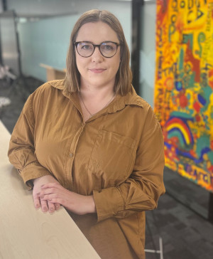 Alyssa Currie, Acting Manager, Procurement and Contracts, Pharmac. She is sitting at a table. Behind her is a bright coloured painting and some glass doors