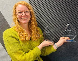 Grace van Dyk holds two awards won this year. Grace has long curly hair, glasses, and is wearing a green top.