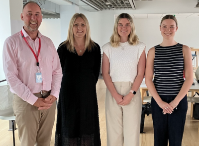 Four people in tidy casual office-wear stand shoulder to shoulder smiling.