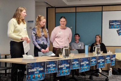 In an office setting, three graduates present at a table decorated with blue Shark Tank 2025 posters, while two of their team look on in support behind them.