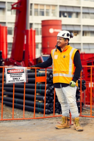A smiling intern wearing a hard-hat and orange high-vis vest is standing at a construction site.