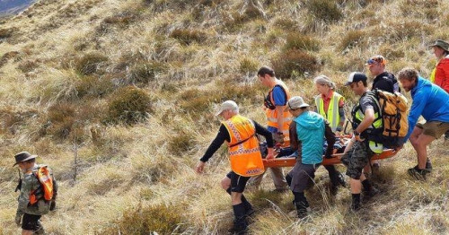 It is a sunny day in the New Zealand wilderness. A group of New Zealand Land Search and Rescue volunteers are carrying an injured person on a stretcher to safety.