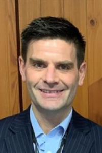 New Zealand Government Procurement General Manager Michael Alp smiles. He is wearing a bright blue shirt and a blue and grey pinstripe jacket.