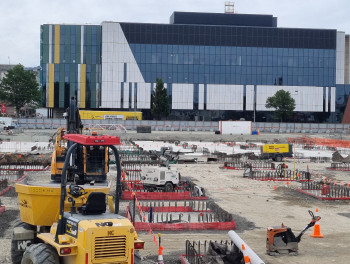 A large building site with yellow machinery and construction in the foreground and a tall windowed building in the background.