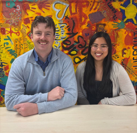 Sam and Mikka are both wearing warm looking tops in this portrait photo. They are standing up and behind them is a bright mural. Both are smiling.