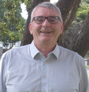 OSPRI Procurement and Contracts Manager Ray Yates. This is a portrait phot. It shows Ray with short cropped hair. He’s wearing glassed and wearing a shirt. He’s in a tree lined street.