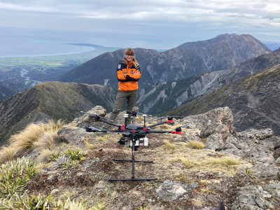 A person in an orange and black jacket with a drone in a mountainous area.