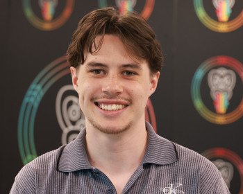 Portrait photo of Tupono with short hair. He’s smiling and wearing a plain shirt. In the background is a panel with promotional material for Māori Language Week.