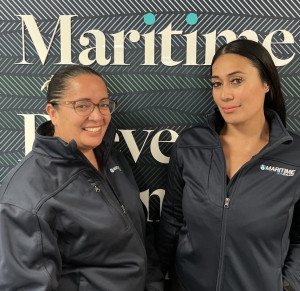 Olivia Tuatoko and Lesa Laga at Maritime NZ. They are both wearing Maritime NZ uniforms which are black. They are standing in front of a Maritime NZ backdrop which is an abstract pattern of waves moving.